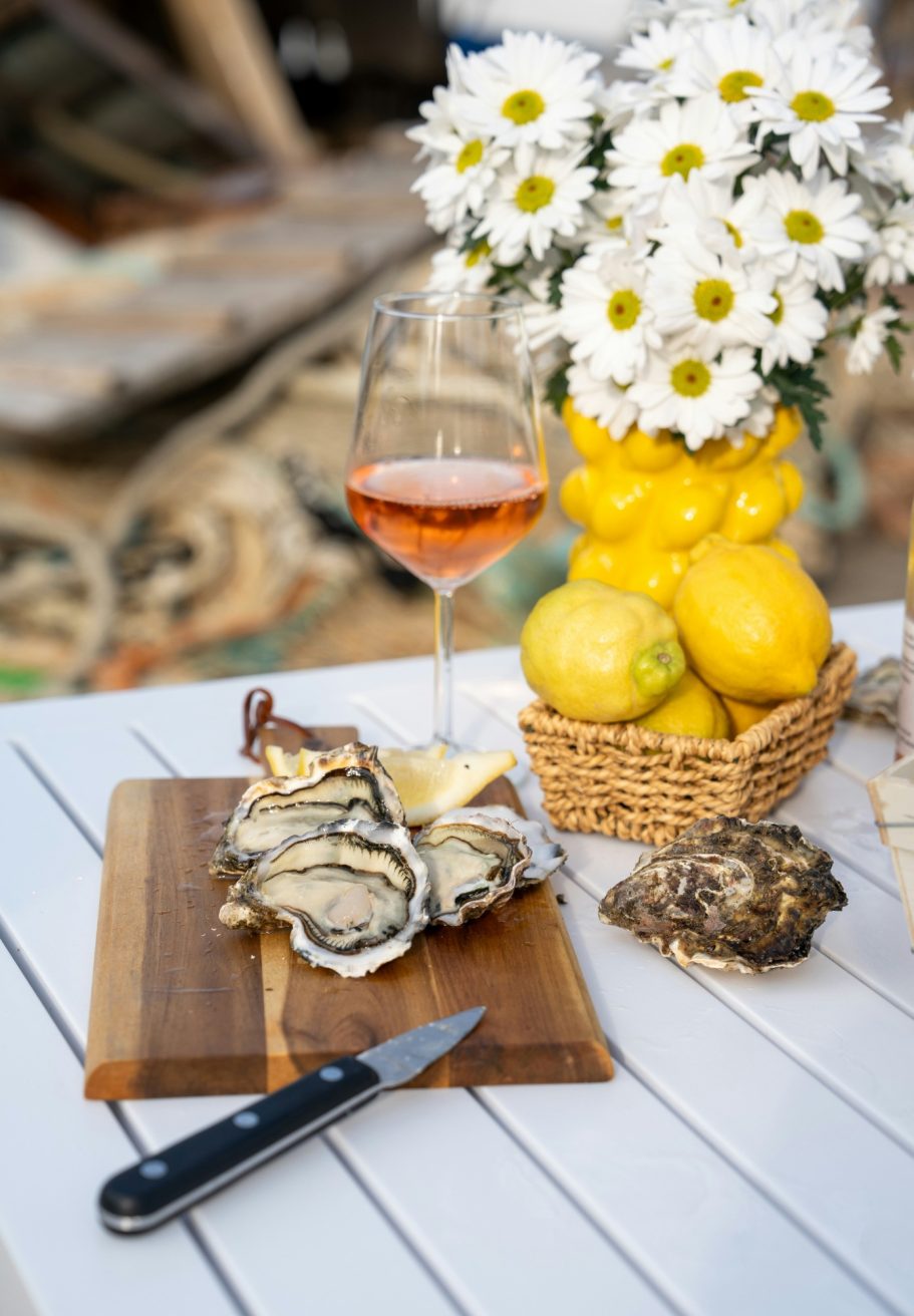 Huîtres sur une planche, verre de rosé, fleurs blanches et citrons en arrière-plan.